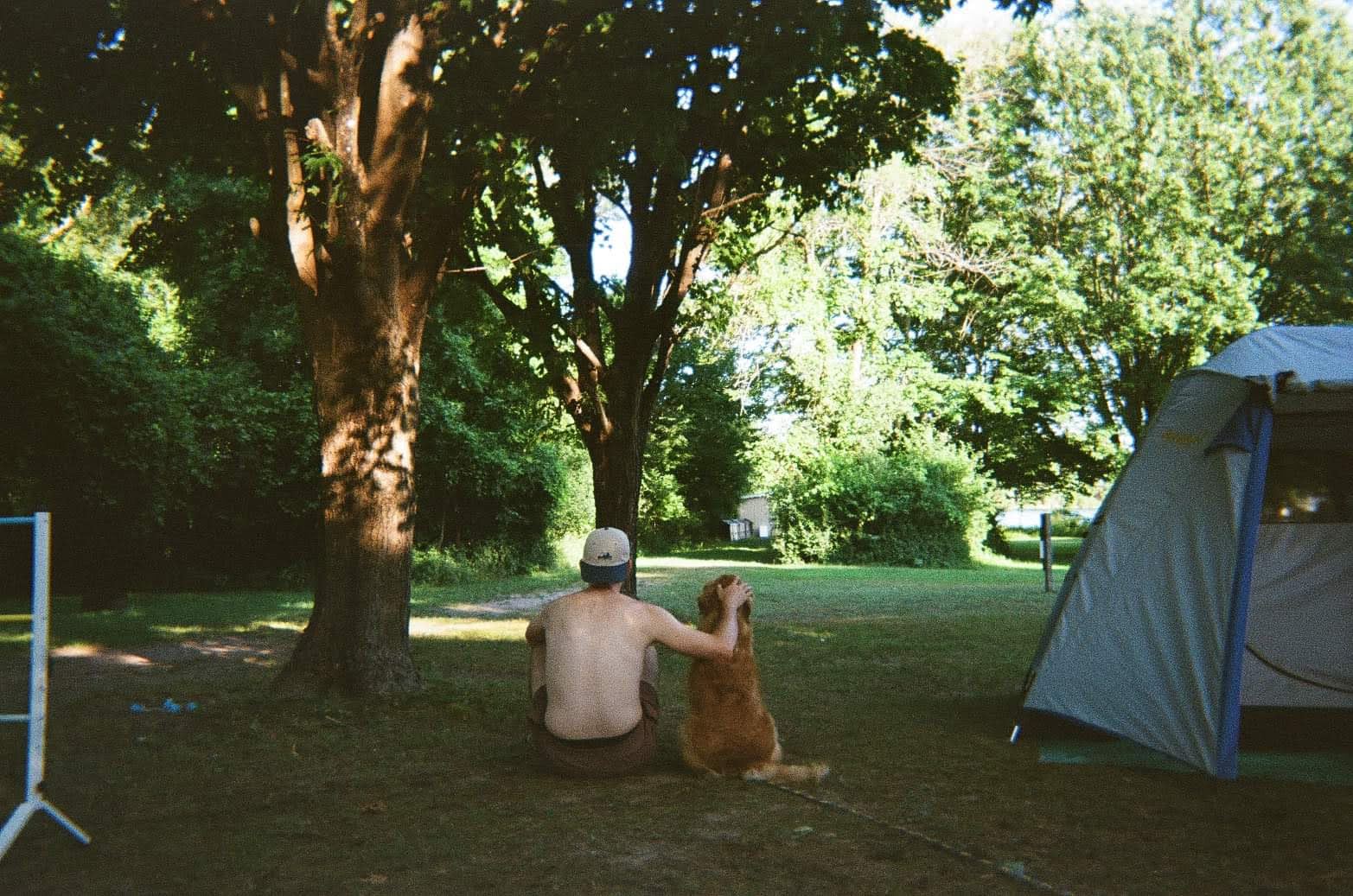 Harrison sitting against a tree at a campsite with a golden dog beside him, a tent visible to the right, shot on film